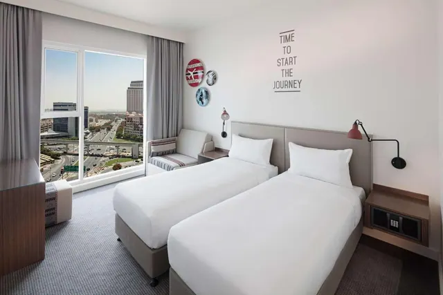 A hotel bedroom with two single beds, a small armchair, and a large window overlooking a road with traffic and city buildings.
