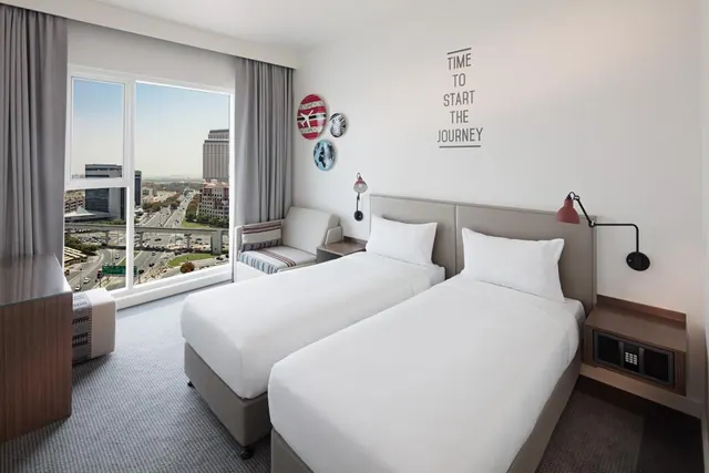 A twin-bedded hotel room with white bedding, a small seating area and a large window overlooking a city highway and high-rise buildings.
