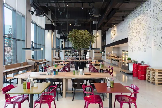 A casual restaurant dining room with wooden tables, red chairs, a central potted tree and a food counter along the right wall.