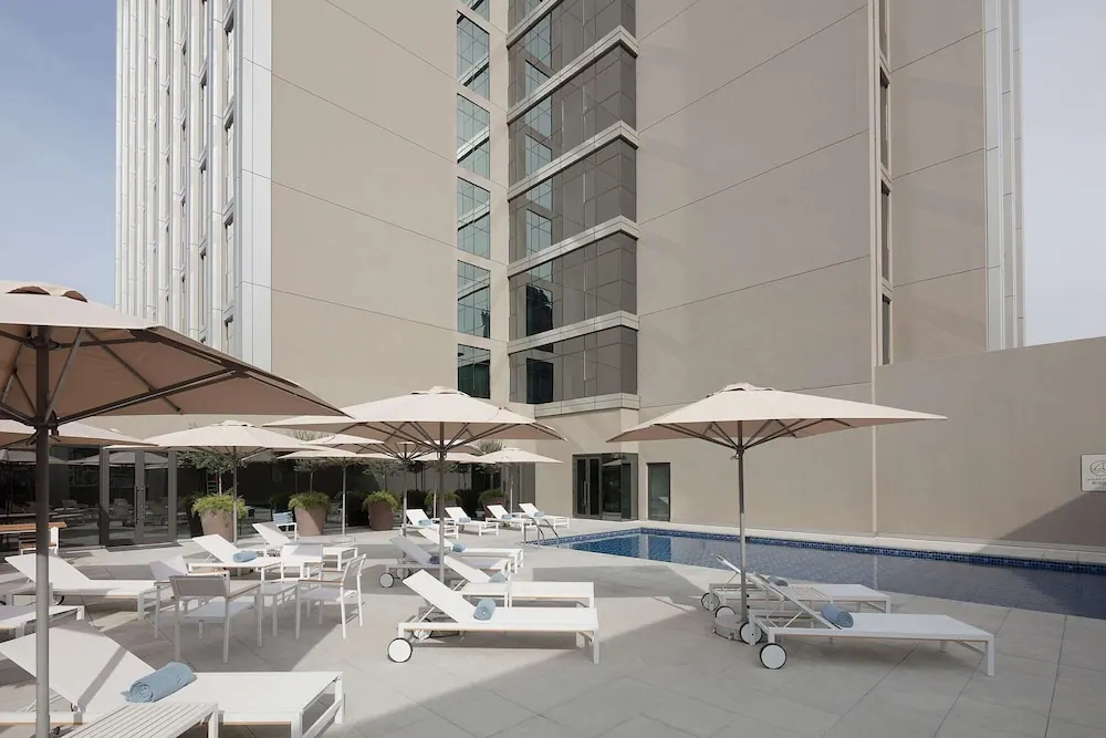 Outdoor pool terrace with white sun loungers, cream shade umbrellas and a tiled pool beside a tall beige hotel building.