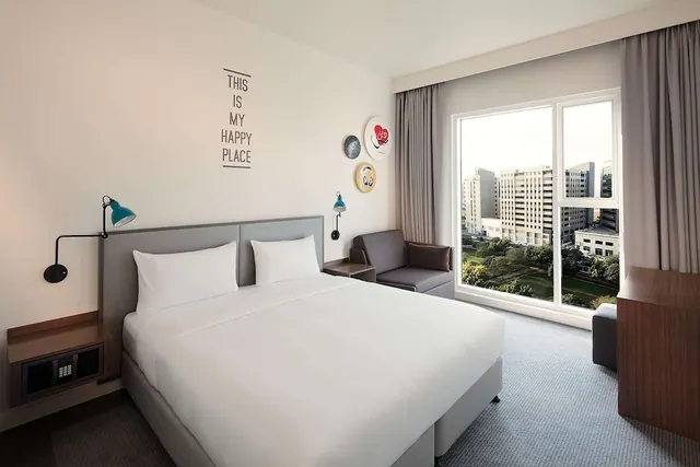 A bedroom with a king bed, a grey armchair, wall lamps and a large window looking out over trees and mid-rise buildings.