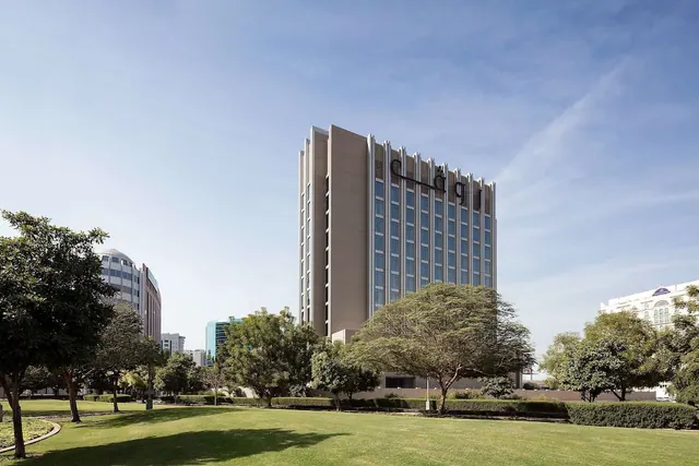 A tall hotel tower with an Arabic sign on its facade stands behind a grassy lawn and trees under a blue sky.