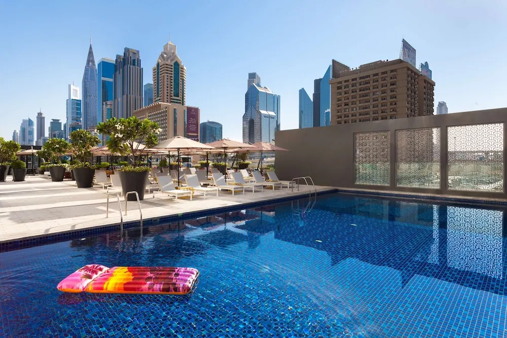 A rooftop pool with blue mosaic tiles, sun loungers and umbrellas, a pink inflatable float in the water, and a city skyline of tall towers behind.
