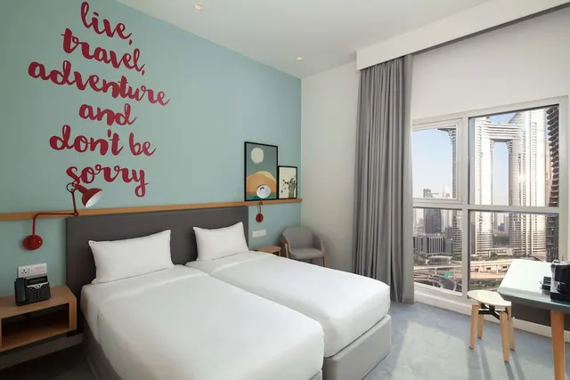 A bedroom with two single beds against a teal wall with red lettering, a seating chair, and a large window overlooking high-rise towers, with a desk to the right.