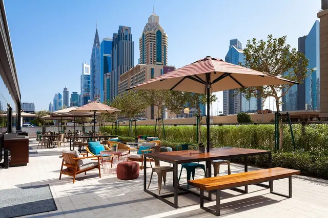 An outdoor terrace with wooden tables, a bench, lounge chairs and striped umbrellas, with a row of high-rise towers beyond the hedge.