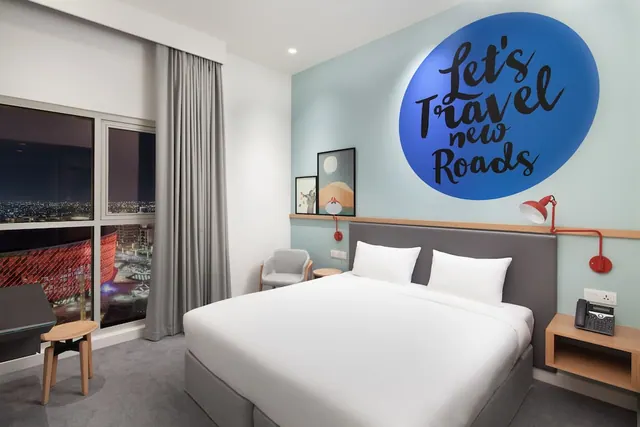 A hotel bedroom with a king bed, an armchair, a large wall graphic reading Let's Travel new Roads, and floor-to-ceiling windows showing a night city view.