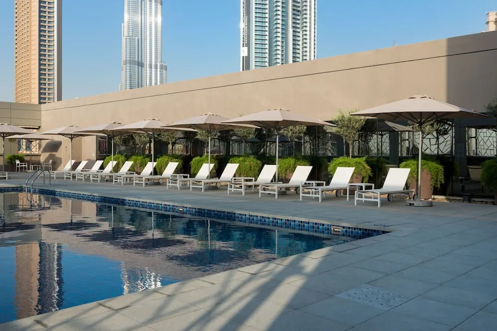 A rectangular swimming pool with white sun loungers and beige umbrellas on the deck, with the Burj Khalifa and other towers rising behind the building.