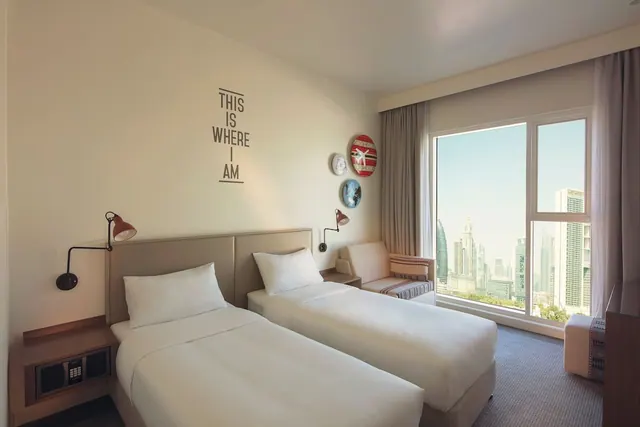 A twin bedroom with two single beds, a seating area by the window, and a view of high-rise towers through floor-to-ceiling glass.
