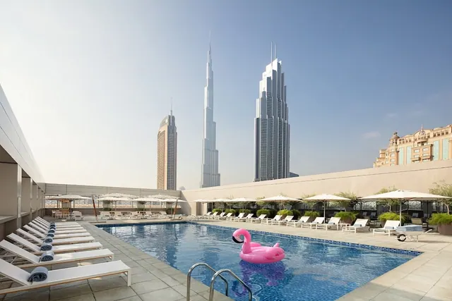 Rooftop pool with white sun loungers and umbrellas and a pink flamingo float in the water, with the Burj Khalifa and other towers visible in the skyline.