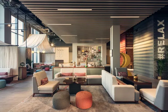 A lounge seating area with light sofas, patterned cushions, knitted poufs and a low coffee table, with a gallery wall and tall windows behind.