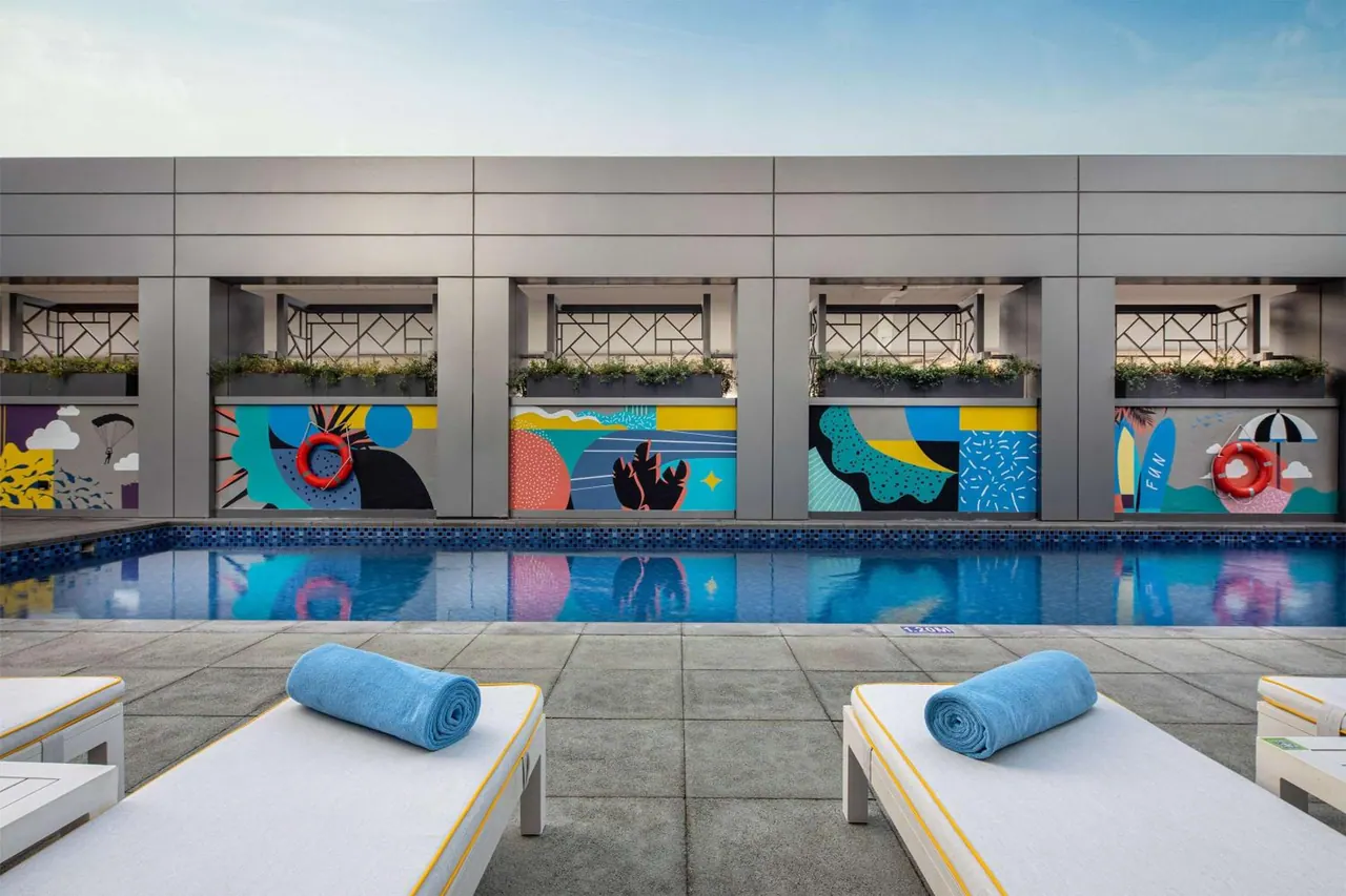 An outdoor pool with colourful mural panels on the wall behind it, and white sun loungers with rolled blue towels in the foreground.