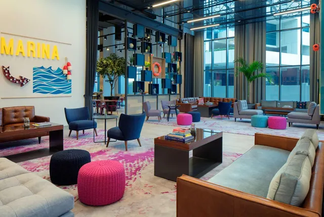 A lobby lounge with leather and grey sofas, low wooden tables, blue armchairs, pink and turquoise poufs, floor-to-ceiling windows and a large yellow wall sign reading MARINA.