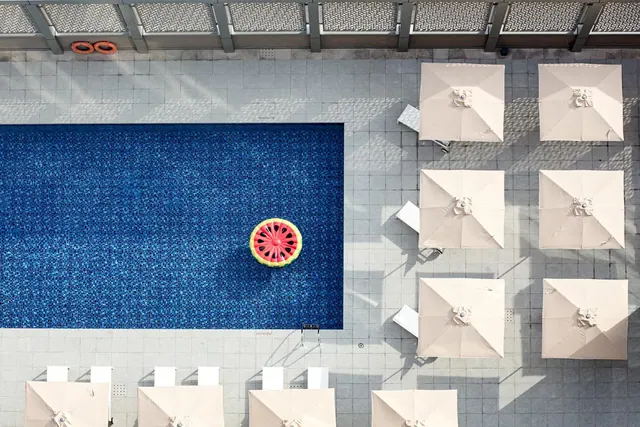 Overhead view of a rectangular swimming pool with a watermelon-shaped float, surrounded by cream square umbrellas and white sun loungers on grey paving.