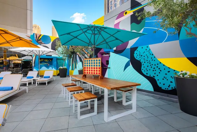 An outdoor terrace with teal and yellow umbrellas, white sun loungers, a wooden table with benches, a giant Connect Four game, and a large colourful mural on the wall.