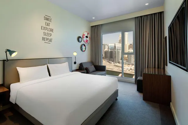 A bedroom with a large white bed, a grey armchair, a wall-mounted television and a sliding window looking out onto city towers and a canal.