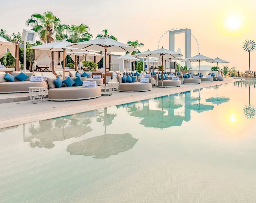 A pool deck with cushioned daybeds, blue cushions and white parasol umbrellas, with the Ain Dubai ferris wheel visible in the background at sunset.