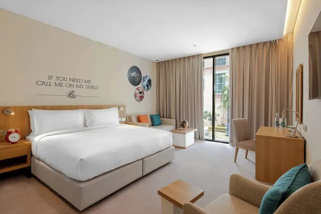 A bedroom with a king bed dressed in white linen, a small sofa with cushions, a wooden desk and chair, and a glass door looking out onto trees and a neighbouring building.