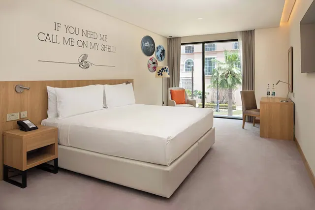A hotel bedroom with a king bed, an armchair with an orange cushion, a wooden desk and chair, and sliding glass doors looking out onto a palm tree and a building.