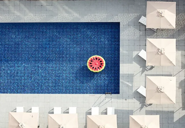 Overhead view of a rectangular blue-tiled pool with a watermelon-shaped inflatable, surrounded by cream umbrellas and white sun loungers.