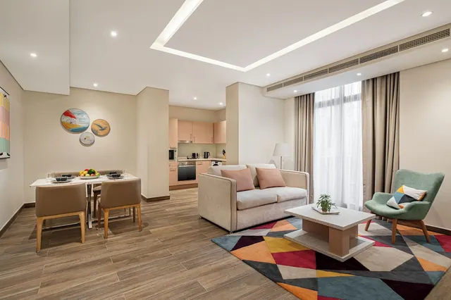 A staged living area with a dining table, a light grey sofa, a multicoloured geometric rug, a green armchair, and an open kitchenette in the background.
