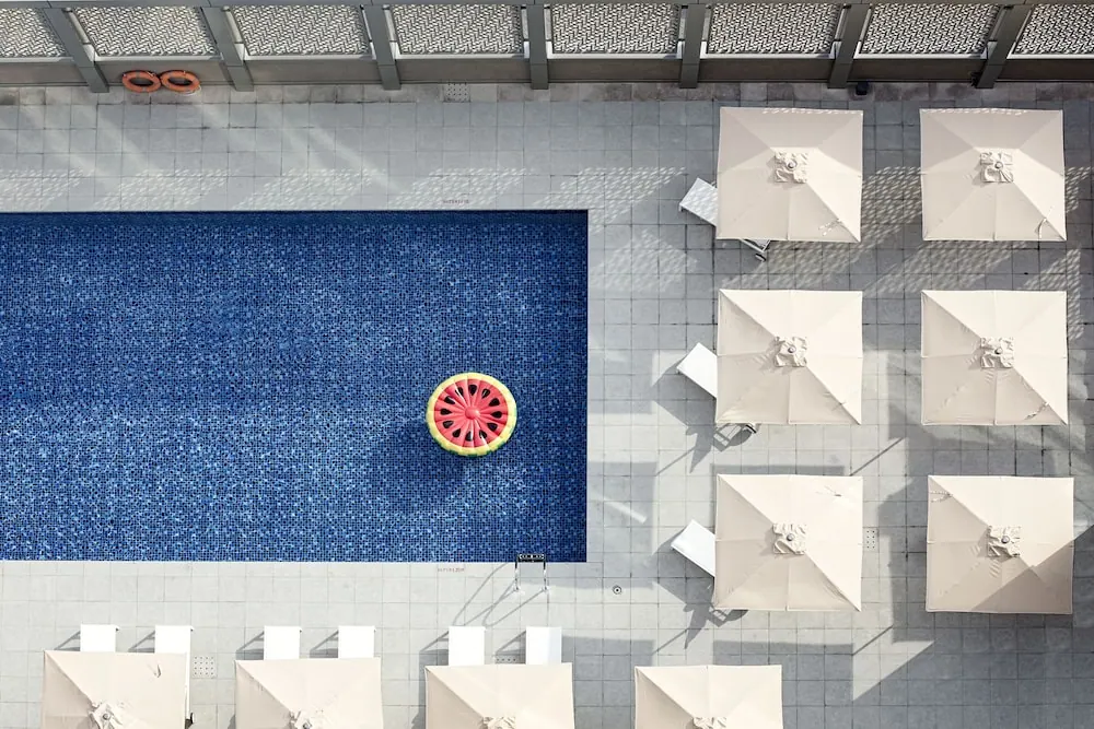Overhead view of a rectangular blue-tiled swimming pool with a watermelon-shaped inflatable float, white umbrellas and sun loungers on the surrounding deck.
