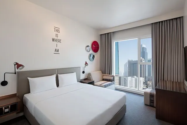 A hotel bedroom with a king bed, a seating area by a large window, and a city skyline of tall buildings visible outside.