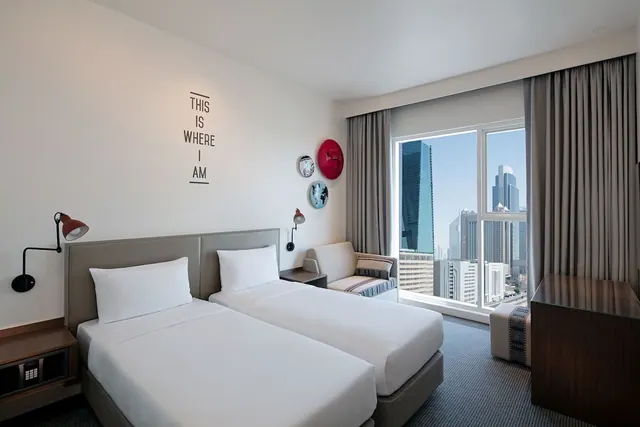 A hotel bedroom with two single beds, a small sofa, and a large window showing high-rise towers outside.
