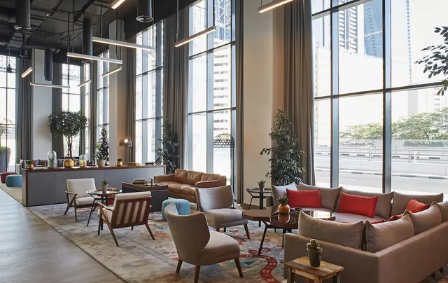 A lobby lounge with beige sofas and armchairs on a patterned rug, with tall windows looking onto city towers and a road outside.