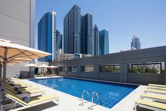 A rooftop swimming pool with yellow-cushioned sun loungers and parasol umbrellas, with tall skyscrapers visible beyond the building edge under a clear blue sky.