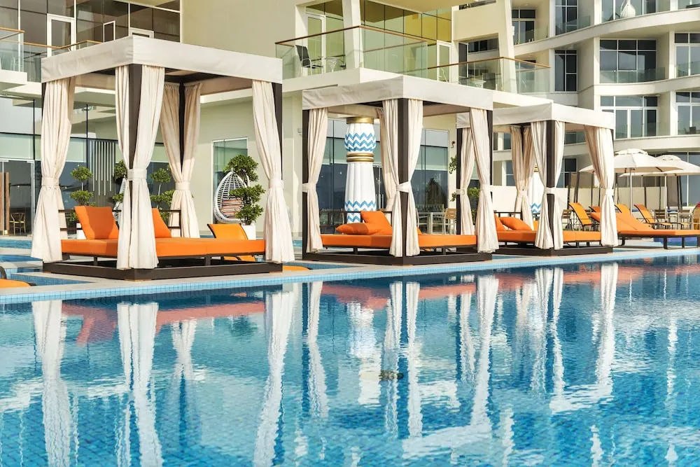 A large swimming pool in front of white canopied cabanas with orange sun loungers, with white parasols visible on the right.