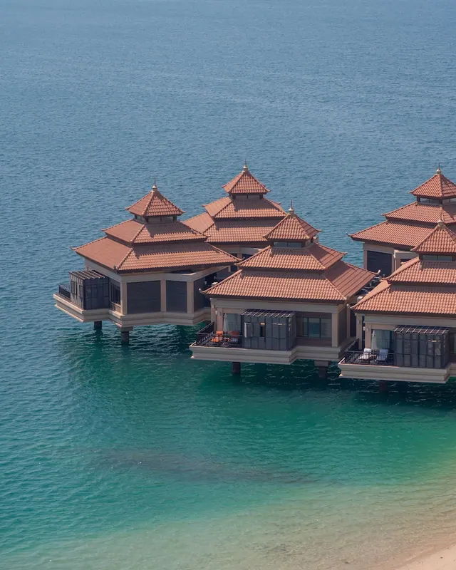 Aerial view of several overwater pavilions with terracotta pagoda-style roofs above turquoise water beside a sandy shore.