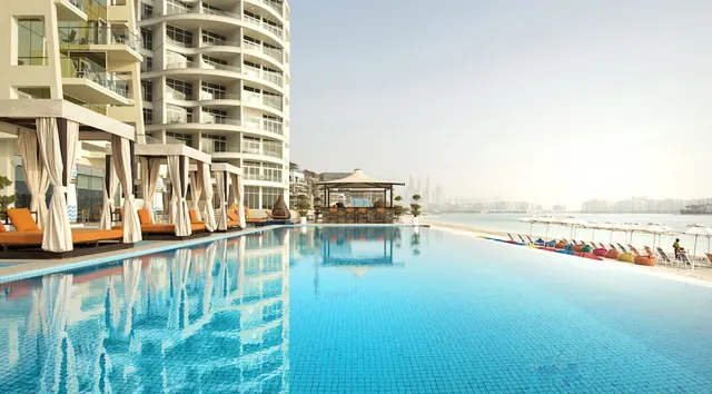 An infinity-edge pool with orange loungers and cabanas beside a tall residential-style building, with a sandy beach, umbrellas and a hazy skyline across the water.