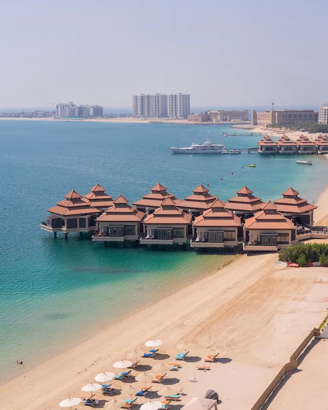 Aerial view of overwater bungalows with terracotta roofs, a sandy beach with sun loungers and umbrellas, and the sea and distant towers beyond.