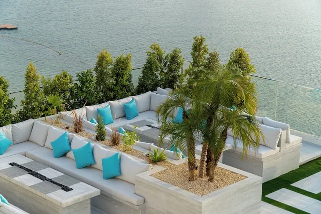 An outdoor terrace with large white sectional sofas, turquoise cushions and palm trees in gravel planters, overlooking calm water behind a glass railing.