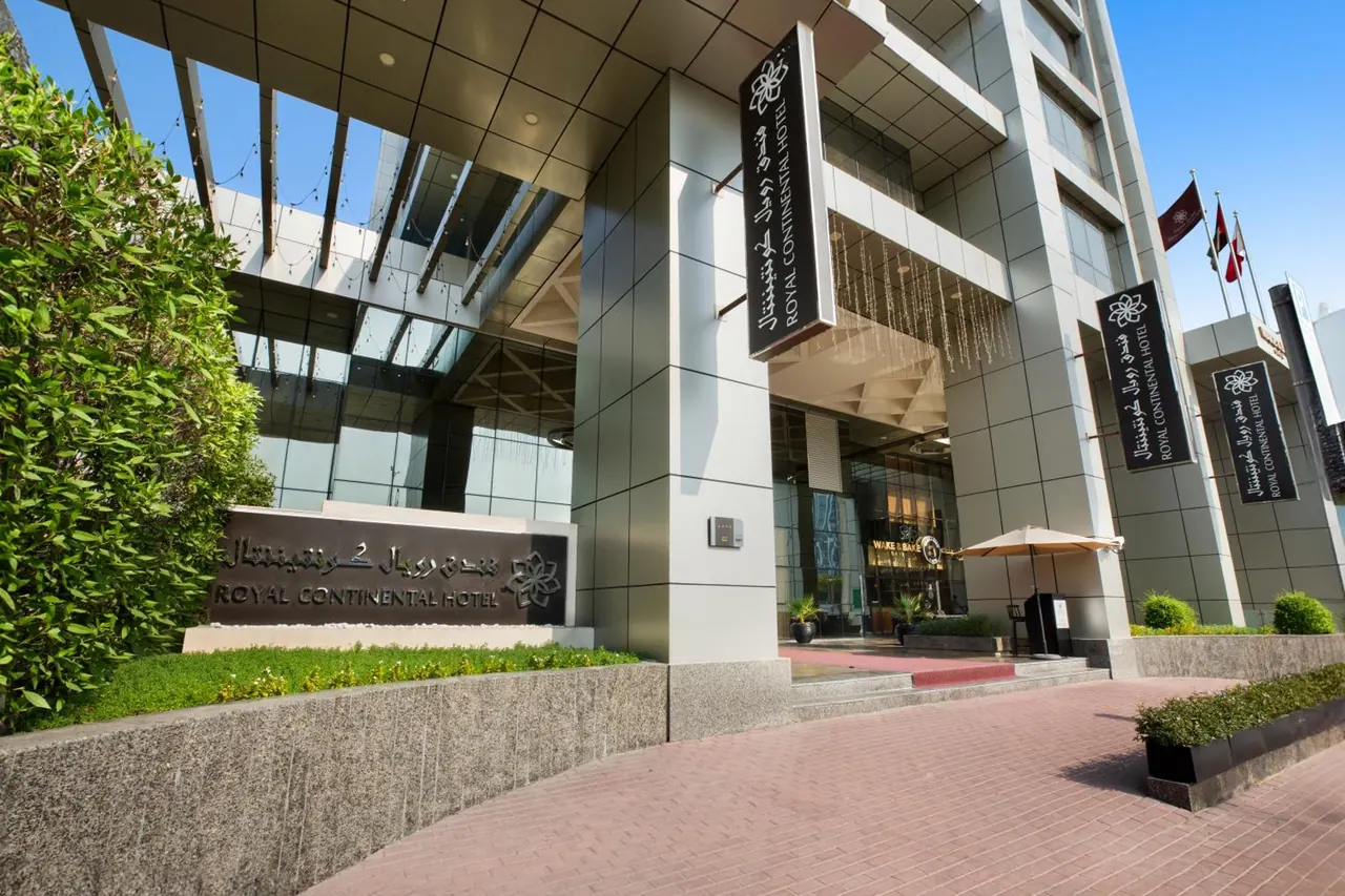 The front entrance of the Royal Continental Hotel, with a stone name sign, vertical banners on the facade, a hedge and a brick paved forecourt under a clear blue sky.