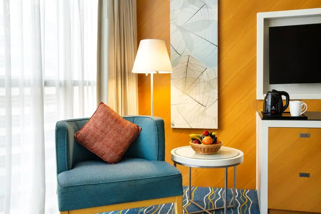 A teal armchair with a red patterned cushion sits beside a floor lamp and a round side table holding a fruit basket, with a television and electric kettle on a cabinet to the right.