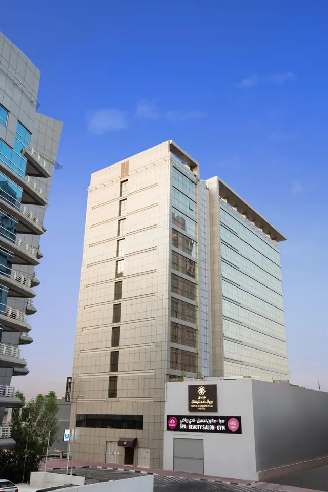 Exterior of a tall hotel tower with a Royal Continental Hotel sign and a spa, beauty salon and gym sign at street level, under a blue sky.