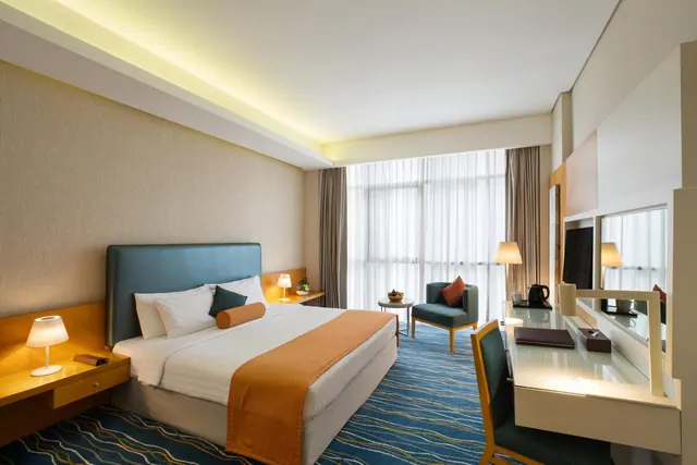 A modern hotel bedroom with a king bed, a teal armchair beside a window with sheer curtains, and a desk with a mirror and kettle along the right wall.