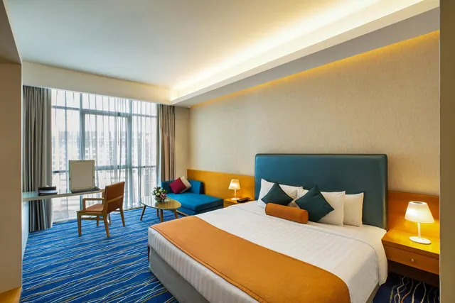 A bedroom with a large bed with a teal headboard, a blue armchair, a desk and chair, and floor-to-ceiling windows looking out over high-rise buildings.
