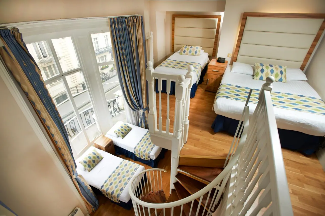 A bedroom with two single beds and a double bed beside a white spiral staircase, with tall windows overlooking a street of buildings.