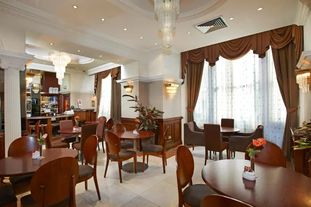 A dining room with round wooden tables and upholstered chairs, a crystal chandelier, draped curtains at tall windows, and a bar in the background.