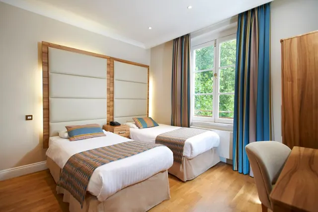 A bedroom with two single beds with upholstered headboards, a wooden wardrobe, a desk with a chair, and a window looking out onto trees.