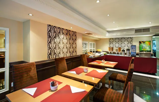 A breakfast dining room with set tables holding red placemats, white napkins and condiment holders, with a juice bar and buffet counter along the back wall.