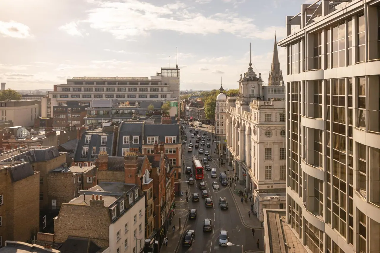 Elevated view down a busy London street with a red double-decker bus, rows of cars, historic stone and brick buildings, and a modern glass office block on the right.