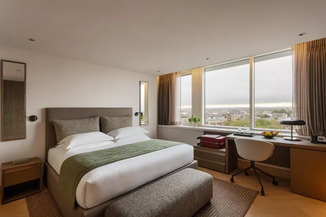 A bedroom with a made double bed, an upholstered bench at its foot, and a wooden desk and chair beside large windows overlooking a city.