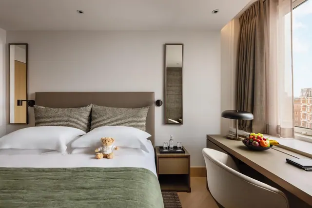 A made double bed with white pillows and a green throw sits beside a desk with a fruit bowl and lamp, next to a window looking out over a brick building.