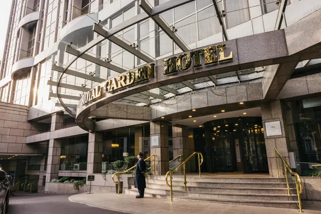 The Royal Garden Hotel entrance shows a glass canopy, gold lettering on the facade, steps with brass handrails, and a uniformed doorman standing at the foot of the steps.