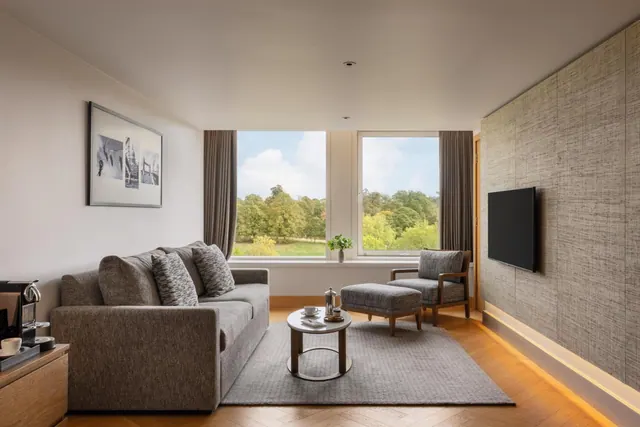 A living area with a grey sofa, armchair, footstool, round coffee table, wall-mounted television and large windows looking onto trees and grass.