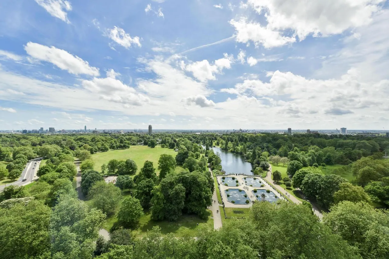 Aerial view over a large green park with a lake, formal fountain gardens and a distant city skyline under a partly cloudy sky.
