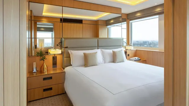 A king bed with white linen sits between wood-panelled walls and mirrored panels, with a window showing a distant city view.